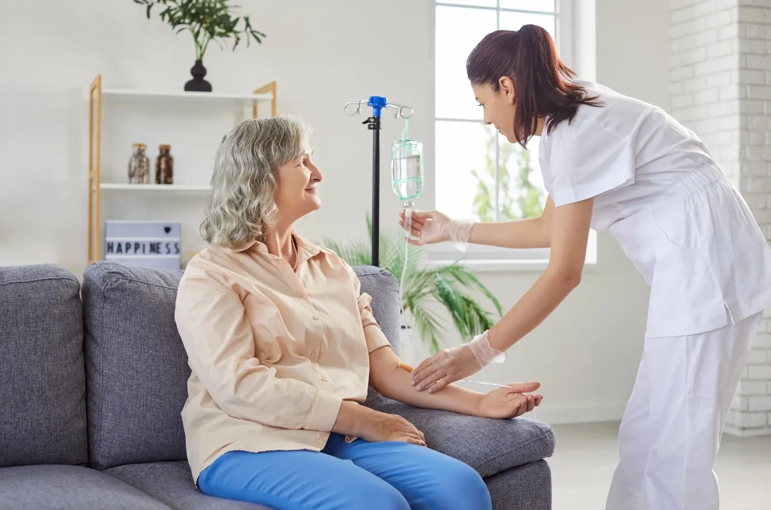 Freundliche junge Krankenschwester hilft einer älteren Patientin, die auf dem Sofa sitzt